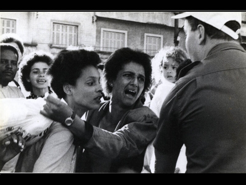 Foto preto-branca de uma mulher gritando/chorando e segurando um(a) jovem observada por 1 criança de colo, 2 jovens e um policial militar de costas