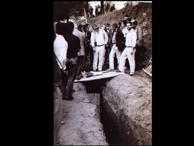 Foto sépia com cerca de vinte homens no entorno de uma vala coberta parcialmente por uma tábua de madeira