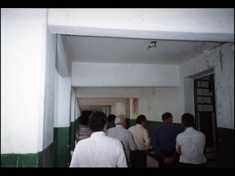 Foto colorida de cinco homens de costas passando por corredor escuro com paredes brancas sujas de infiltração hidráulica