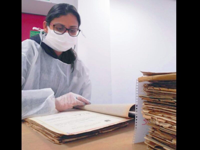 Mulher jovem de cabelos pretos presos, usando óculos e máscara, vestindo avental branco manuseia documento de luvas em frente a uma pilha de outros documentos