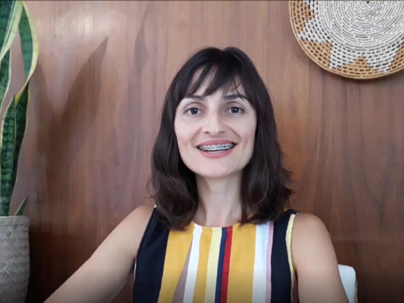 Foto de mulher jovem de pele clara e cabelos escuros, sorrindo sobre parede amadeiradas,