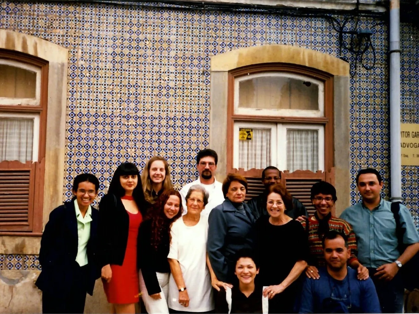 Grupo de 13 adultos reunidos em torno de uma senhora de cabelos escuros e óculos, do lado externo de uma casa com revestimento de ladrilhos portugueses e 2 janelas