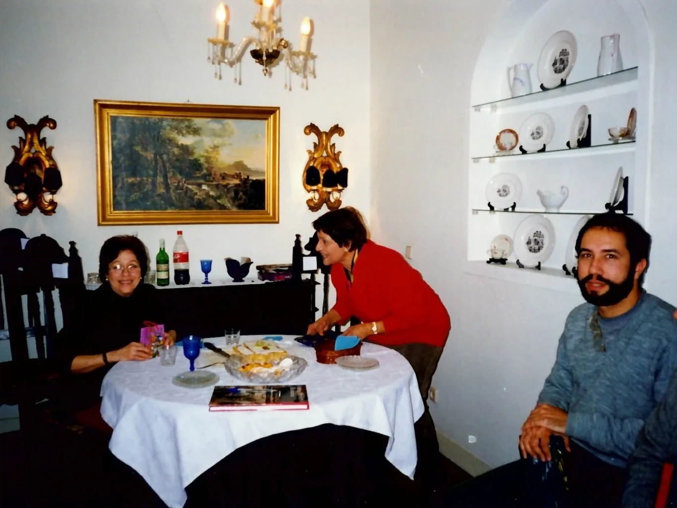 duas senhora de cabelos escuros em torno de mesa com sobremesa, ao fundo garrafas de refrigerante e vinho sobre bancada e quadro na parede, ao lado um rapaz de barba sentado à frente de um painel de nicho de porcelana expostos