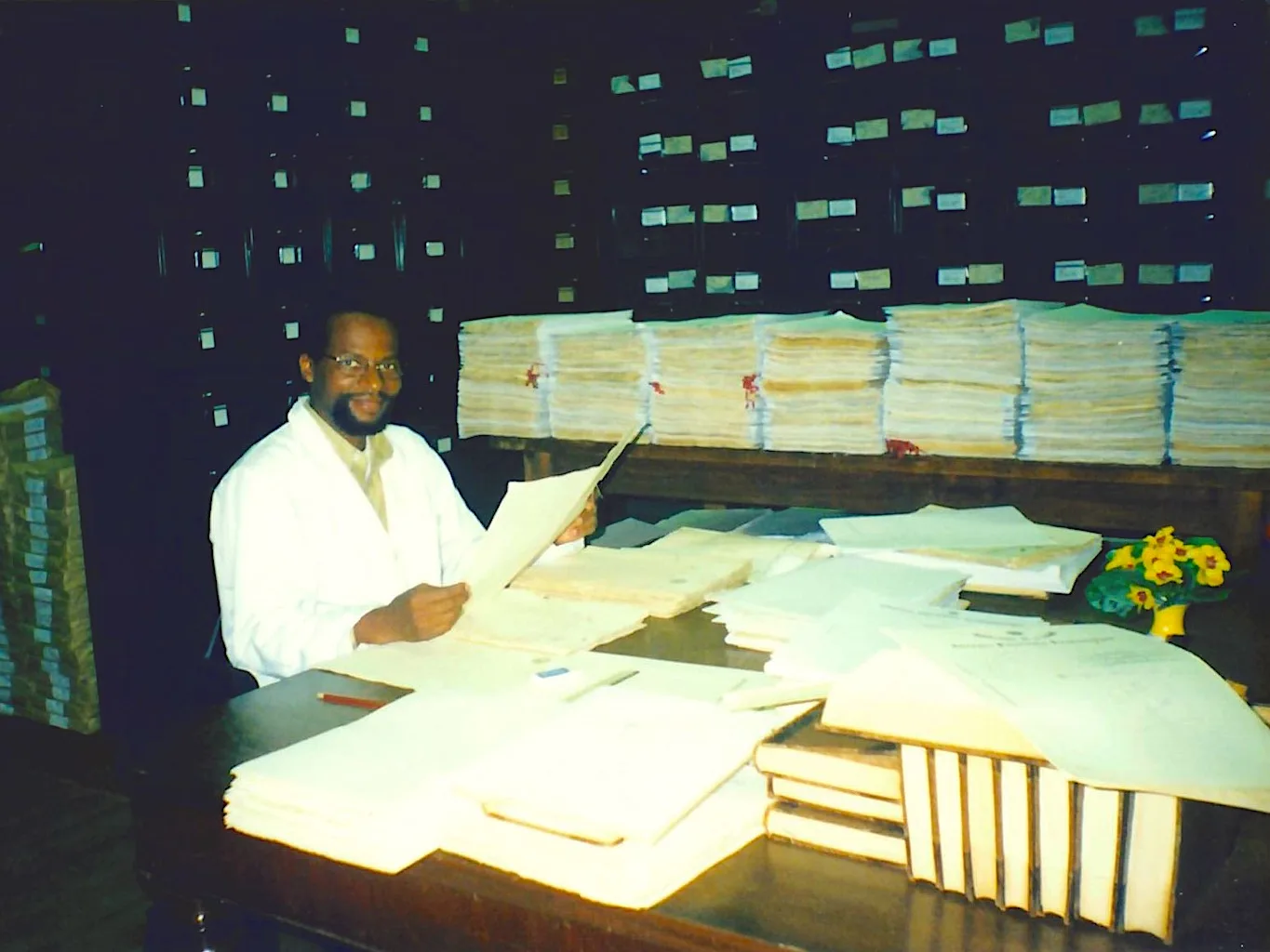 Um homem negro de óculos e cavanhaque, vestido de avental branco, segura documento sobre mesa repleta de outros documentos, ao fundo gaveteiros de madeira escura coom adesivos brancos