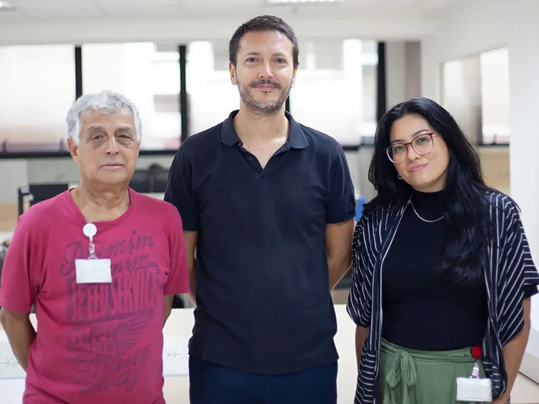 Foto de homem branco de cerca de 40 anos, vestindo camisa preta, posa entre um senhor grisalho de camiseta vermelha e uma mulher de cabelo liso escuro e óculos com blusa escura, com uma janela ao fundo