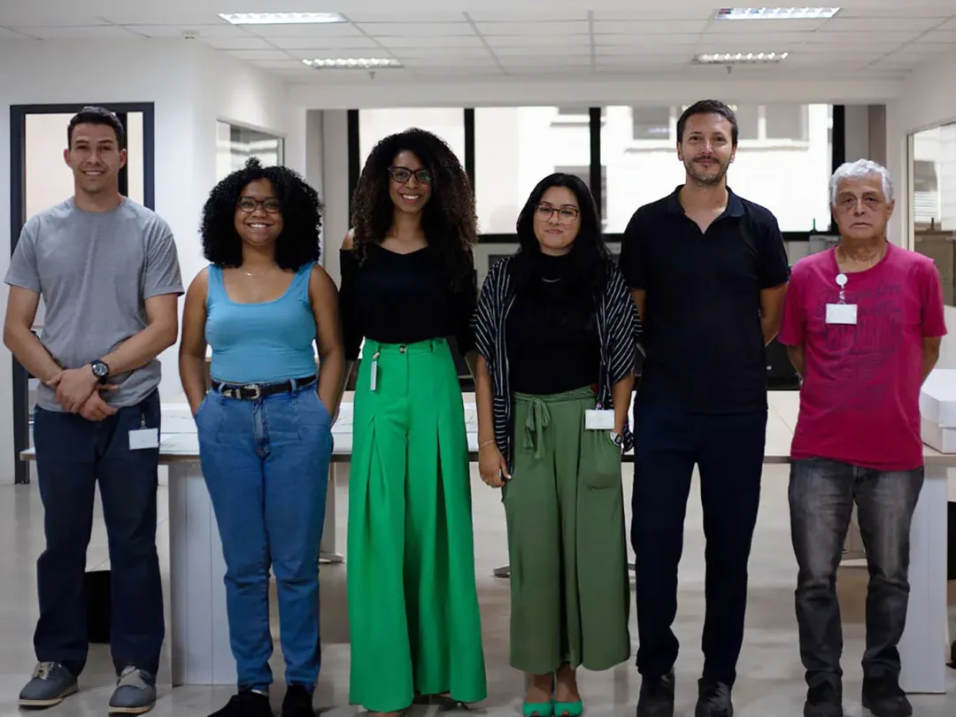 Foto posada de 5 colaboradores perfiladas no entorno do pesquisador, tendo uma mesa e uma janela atrás de si