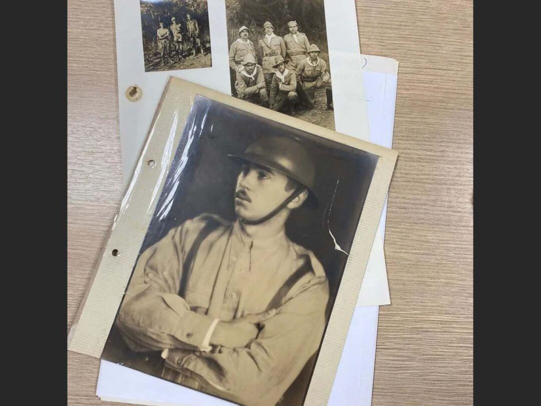 Fotografia em preto-e-branco amarelada de retrato de um homem branco jovem com uniforme de soldado, sobre mesa com outras fotos ao fundo