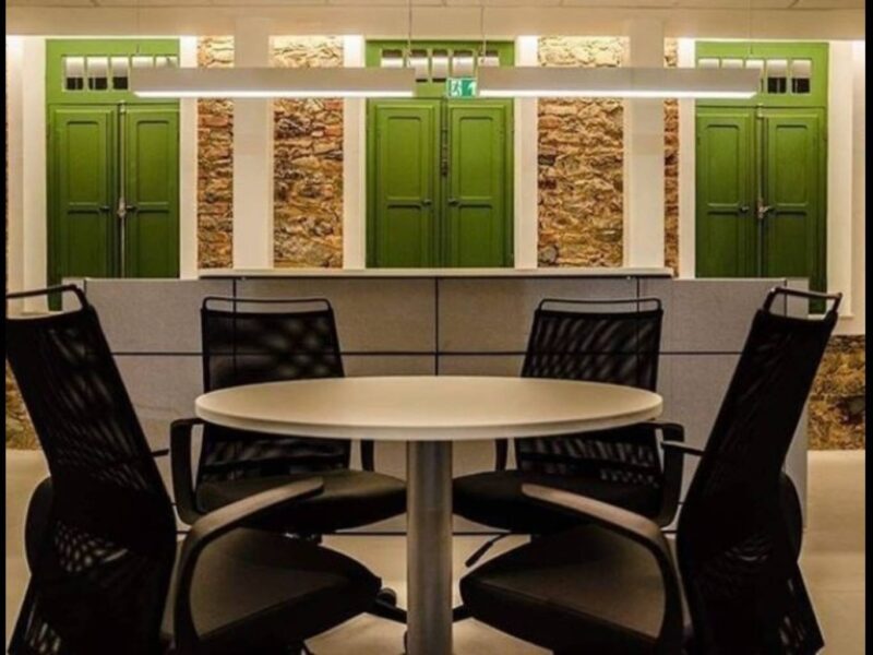 Foto de mesa redonda com quatro cadeiras com encostos de couro preto sob luminária acesa, com parede rústica e portas verdes ao fundo